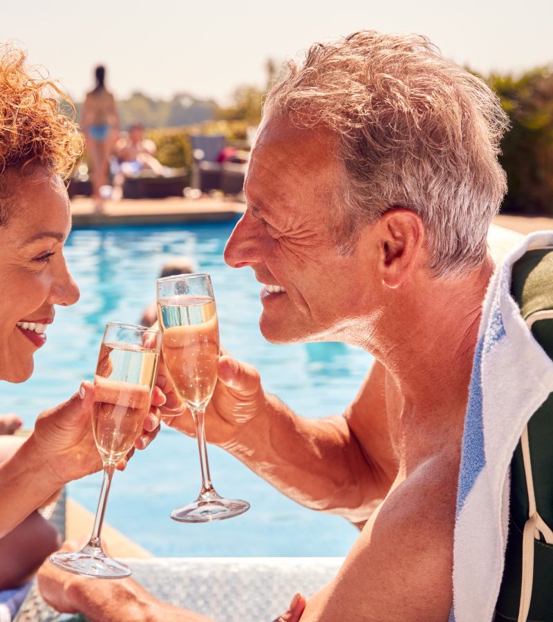 Retired Senior Couple On Loungers Relaxing By Swimming Pool On Summer Vacation Drinking Champagne